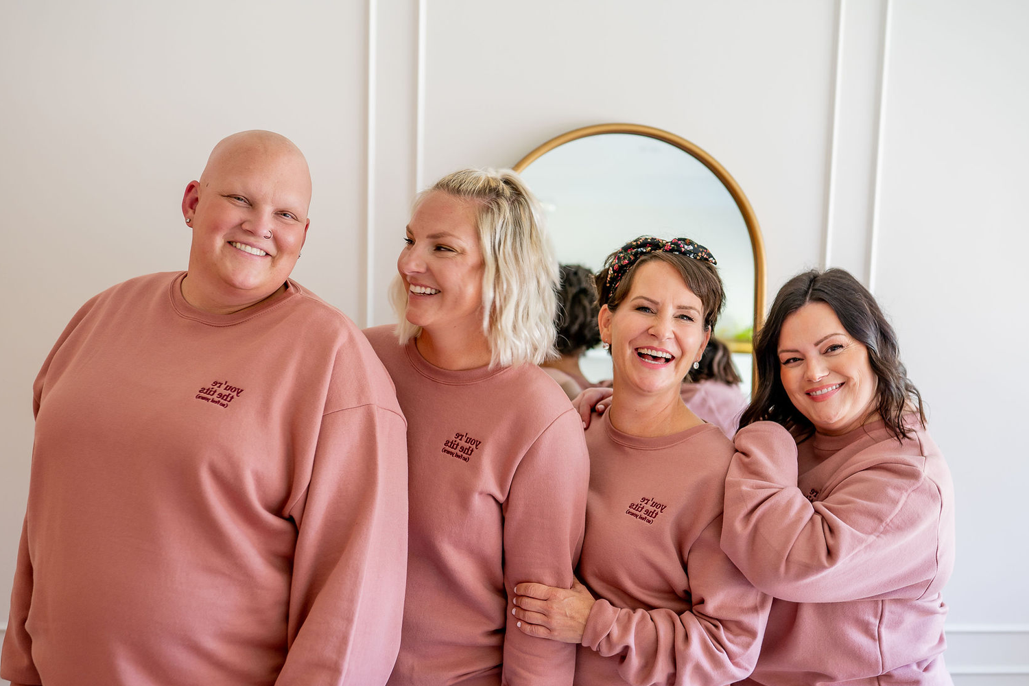 Four women with breast cancer wearing sweaters that say 'you're the tits (so feel yours)"